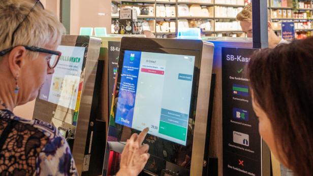Zwei Personen bedienen eine Selbstbedienungskasse in einem modernen Geschäft mit Regalen im Hintergrund.