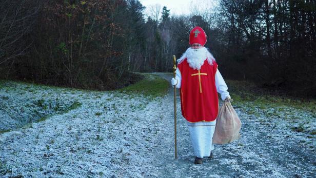 Eine als Nikolaus verkleidete Person mit rotem Mantel, Bischofsmütze und Stab geht auf einem frostigen Waldweg.