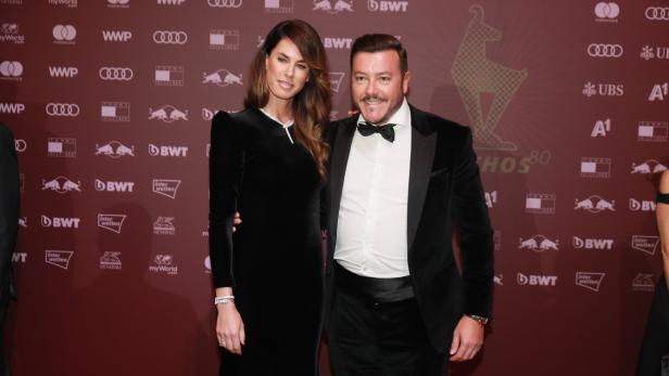 Ein elegant gekleidetes Paar posiert lächelnd vor einer rotbraunen Wand mit Sponsorenlogos.