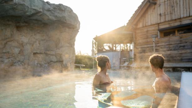 Zwei Personen entspannen im dampfenden Außenpool vor einer Holzhütte bei Sonnenuntergang.