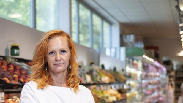 Eine Frau mit roten Haaren steht mit verschränkten Armen vor einer Auslage mit Obst und Gemüse in einem Supermarkt.