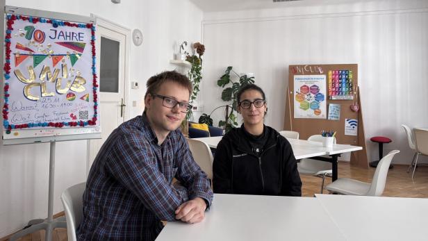 Zwei Personen sitzen an einem Tisch in einem hellen Raum mit bunten Plakaten und Pflanzen im Hintergrund.