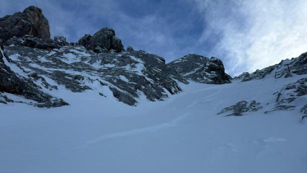 Verschneite Felswand mit steilem Anstieg, im Vordergrund ein Sicherungsseil, darüber bewölkter Himmel.