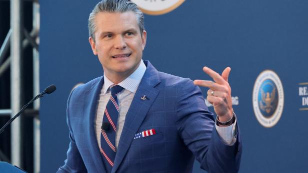Pete Hegseth gestikuliert vor einem blauen Hintergrund mit Logos.
