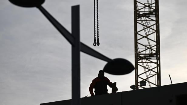 Eine Person arbeitet auf einer Baustelle neben einem Kran, im Vordergrund sind Straßenlaternen zu sehen.