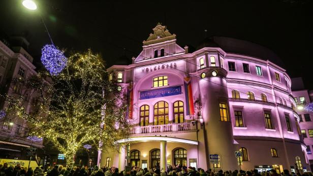 Die Wiener Volksoper erstrahlt nachts in hellem Licht, davor eine Menschenmenge und ein beleuchteter Baum.