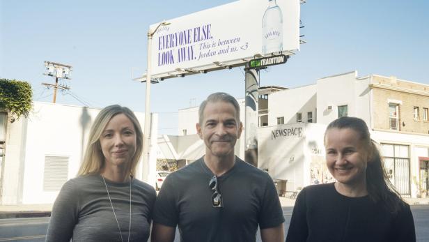 Drei Personen stehen vor einer Werbetafel für Vöslauer Mineralwasser.