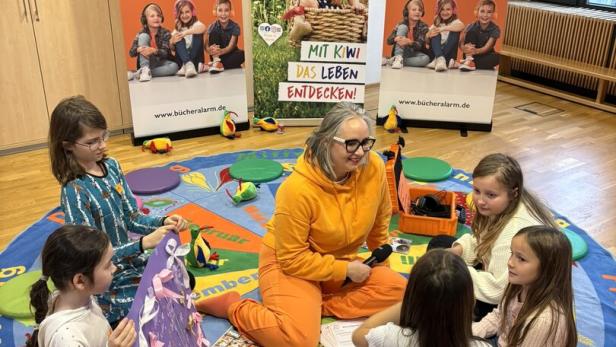 Eine Frau in Orange sitzt mit Kindern auf einem Teppich vor Bannern von "Bücher Alarm" und "Kinder in Wien".