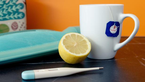 Tasse mit Teebeutel, halbe Zitrone, Wärmflasche und Fieberthermometer auf Tisch.