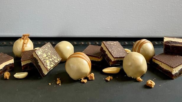 Mehrere Pralinen und Schokoladenstücke mit goldenen Verzierungen und Erdnüssen liegen auf einer dunklen Oberfläche.