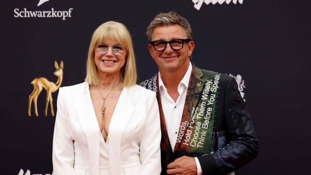 Eine Frau in weißem Anzug und ein Mann mit bedrucktem Sakko stehen lächelnd nebeneinander vor einer dunklen Wand mit Logos.