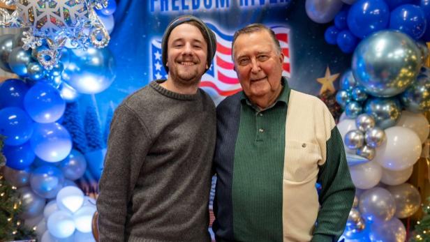 Zwei Männer lächeln vor einer festlichen Ballon-Dekoration mit US-Flagge im Hintergrund.