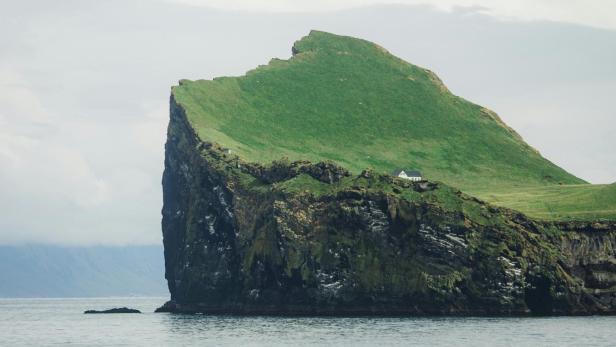 Weißes Haus steht einsam auf grüner Klippeninsel im Meer unter bewölktem Himmel.