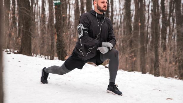 Ein Mann macht einen Ausfallschritt im verschneiten Wald und trägt Sportkleidung sowie Kopfhörer.
