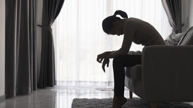 Eine Frau sitzt mit gesenktem Kopf und hängenden Schultern auf einem Sofa in einem abgedunkelten Raum.