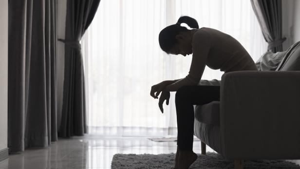 Eine Frau sitzt mit gesenktem Kopf und hängenden Schultern auf einem Sofa in einem abgedunkelten Raum.