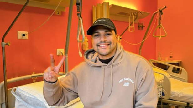 Infleuncer sitzt auf einem Krankenbett und hebt einen Daumen nach oben. 