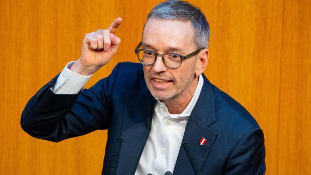 Ein Mann mit Brille gestikuliert vor einem orangefarbenen Hintergrund mit Mikrofonen.
