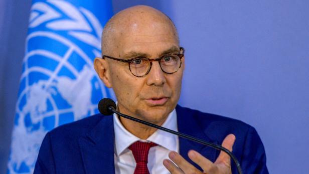 Ein Mann mit Brille und Anzug spricht vor einem Mikrofon, im Hintergrund ist eine UN-Flagge zu sehen.