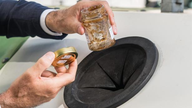 Eine männliche Hand hält ein leeres Marmeladeglas vor einem Glascontainer.