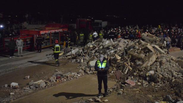 Rettungskräfte und viele Menschen stehen nachts vor einem großen Trümmerhaufen aus eingestürzten Gebäuden.