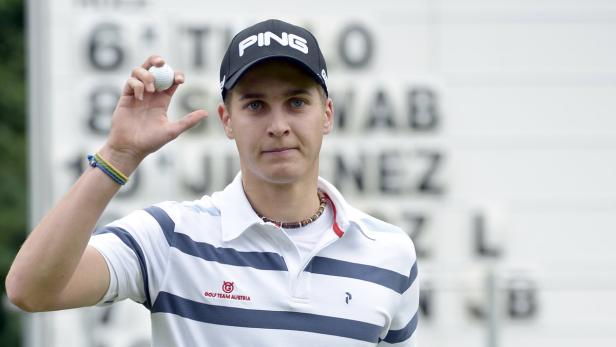 Ein junger Mann mit einer Ping-Kappe hält einen Golfball hoch.