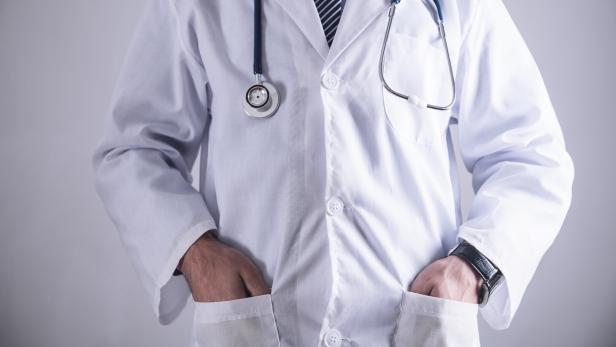 Ein Arzt im weißen Kittel mit Stethoskop und gestreifter Krawatte steht mit den Händen in den Taschen da.