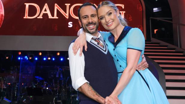 Ein Mann und eine Frau in eleganter Kleidung umarmen sich lächelnd auf einer Bühne mit Lichtern im Hintergrund.