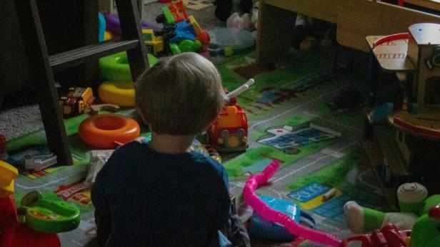 Ein kleines Kind sitzt in einem unaufgeräumten Kinderzimmer voller Spielzeug auf dem Boden.