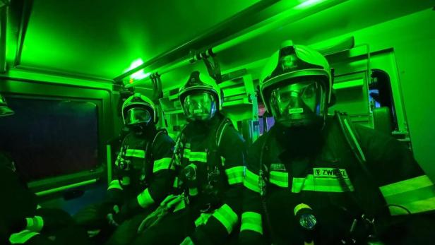 Drei Feuerwehrleute in voller Schutzausrüstung sitzen in einem grün beleuchteten Einsatzfahrzeug.