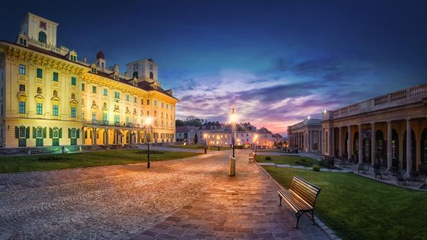 Historisches Schloss mit beleuchteter Fassade, Kopfsteinpflasterweg, Bänken und gepflegtem Garten bei Sonnenuntergang.