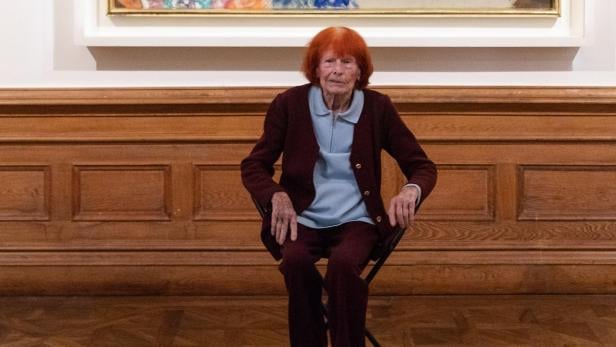 Eine ältere Frau mit roten Haaren sitzt auf einem Stuhl vor einem farbenfrohen Gemälde mit mehreren Figuren.