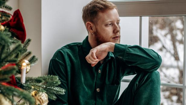 Ein Mann in grüner Kleidung sitzt nachdenklich am Fenster, neben ihm steht ein geschmückter Weihnachtsbaum.