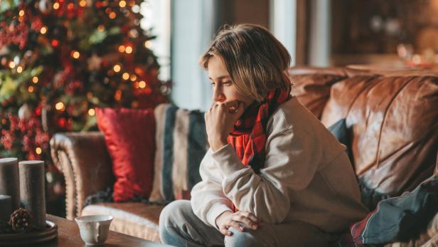Eine nachdenkliche Person sitzt auf einem Sofa mit Schal, im Hintergrund steht ein geschmückter Weihnachtsbaum.