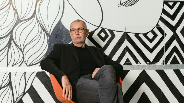 Konrad Paul Liessmann sitzt entspannt auf einem Stuhl vor einer schwarz-weiß gestalteten Wand mit abstrakten Mustern und Zeichnungen.