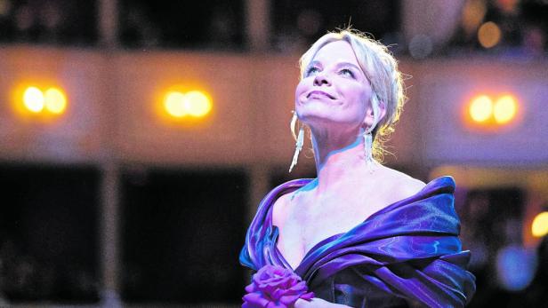 Eine elegant gekleidete Frau in einem lila Kleid hält eine Blume und blickt lächelnd nach oben in einem festlich beleuchteten Saal.