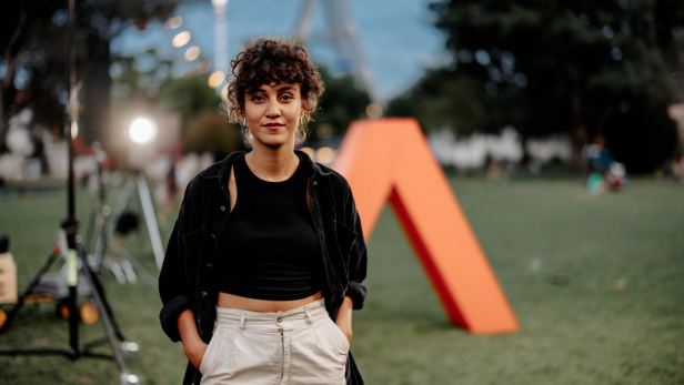 Junge Frau steht auf einer Wiese vor einer großen orangen Skulptur in A-Form.