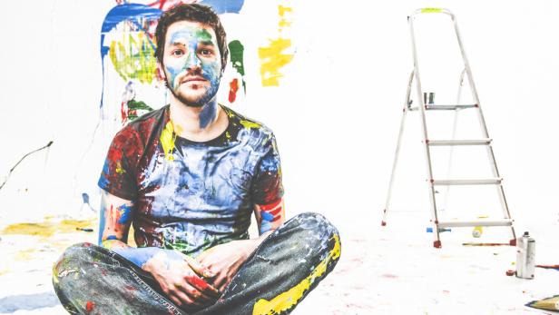 Eine farbige Darstellung eines Mannes, der nachdenklich am Boden sitzt, hinter ihm eine Wand mit Farbklecksen.