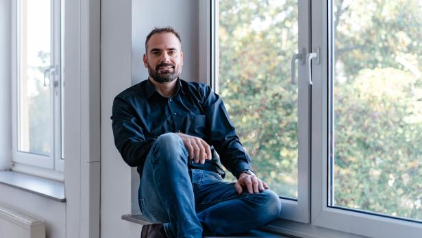 Mann mit Bart sitzt entspannt auf einer Fensterbank vor einem großen Fenster mit Blick ins Grüne.
