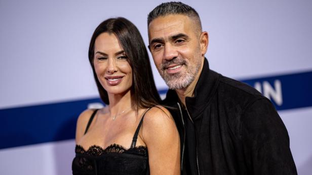 Eine Frau mit dunklem Haar und ein Mann mit Bart lächeln vor einem hellen Hintergrund.