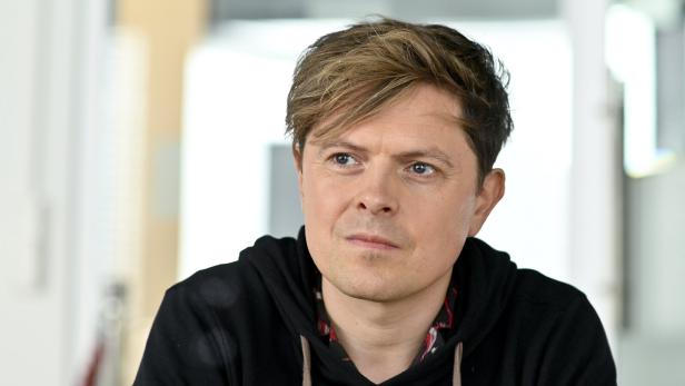 Ein Mann mit blond-braunen Haaren trägt einen schwarzen Hoodie und blickt nachdenklich zur Seite.