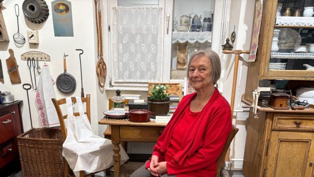 Eine ältere Frau mit grauen Haaren und rotem Oberteil sitzt in einer gemütlichen, altmodischen Küche auf einem Stuhl.