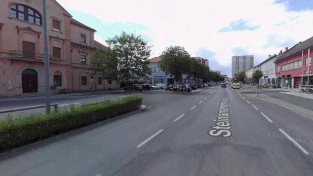 Breite Straße mit mehreren Fahrspuren, flankiert von Bäumen, Autos und mehrstöckigen Gebäuden auf beiden Seiten.