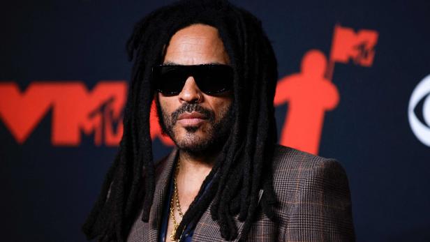 Eine Person mit langen Dreadlocks, Sonnenbrille und kariertem Sakko steht vor einem schwarzen Hintergrund mit roten Symbolen.