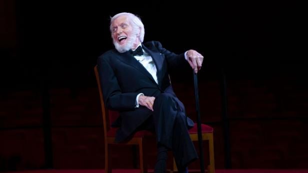 Ein älterer Mann im Smoking sitzt lachend auf einem Stuhl und hält einen Gehstock auf einer Bühne.