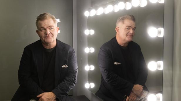 Ein Mann sitzt vor einem beleuchteten Spiegel und schaut nachdenklich, sein Spiegelbild ist deutlich zu sehen.