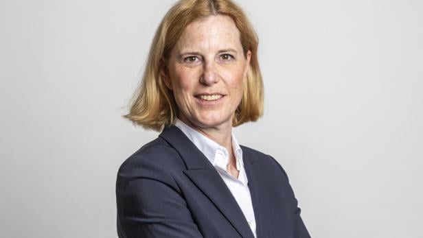 Frau mit schulterlangem blondem Haar trägt einen dunklen Anzug und steht mit verschränkten Armen vor hellem Hintergrund.