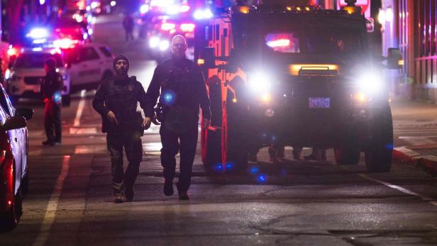 Zwei Polizisten in Schutzkleidung gehen nachts vor einem gepanzerten Fahrzeug mit eingeschaltetem Blaulicht auf einer Straße.