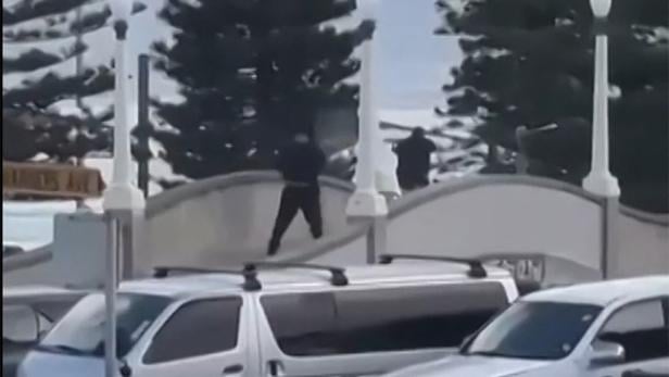 Drei Personen in dunkler Kleidung stehen auf einer Brücke, im Vordergrund parken mehrere weiße Autos.