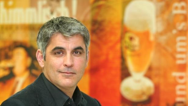 Ein Mann mit grau meliertem Haar steht vor einem Werbeplakat für Bier.